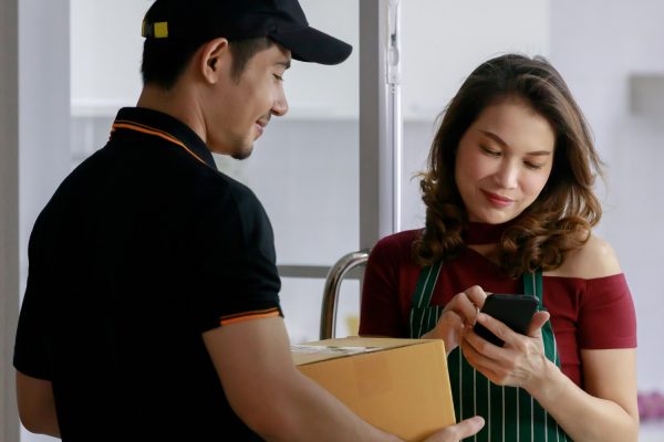 handsome-asian-courier-man-delivers-postal-package-to-attractive-asian-female-shop-owner-who-signs-electronic-signature-on-mobile-device-parcel-shipping-e-commerce-business-concept-1